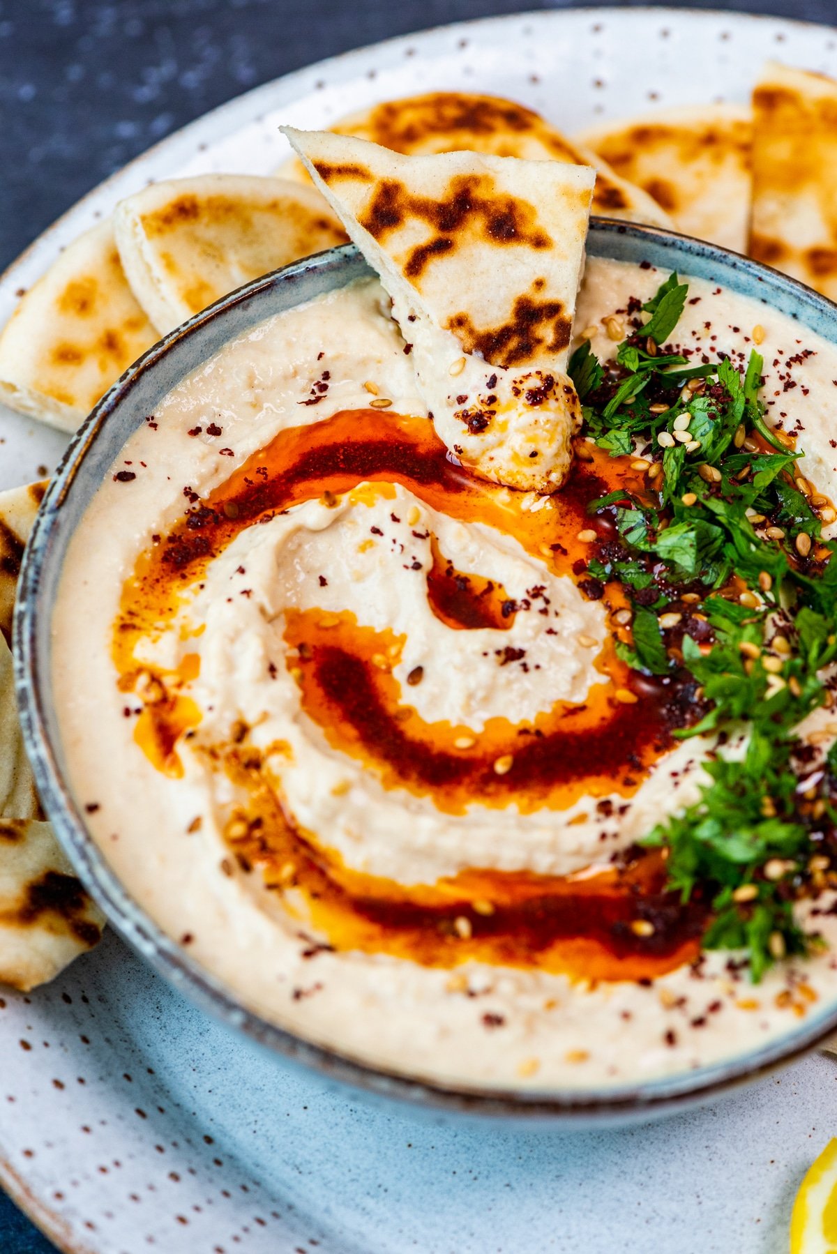 Creamy hummus dip in a bowl garnished with paprika oil, parsley, sumac and sesame seeds and a piece of pide bread in it.