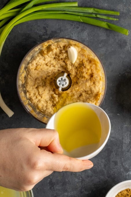 Z hand pouring olive oil from a small white bowl over mashed chickpeas in a food processor.