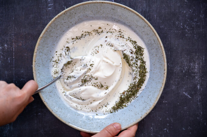 Hands mixing yogurt, dried mint and salt in a large blueish bowl with a spoon.