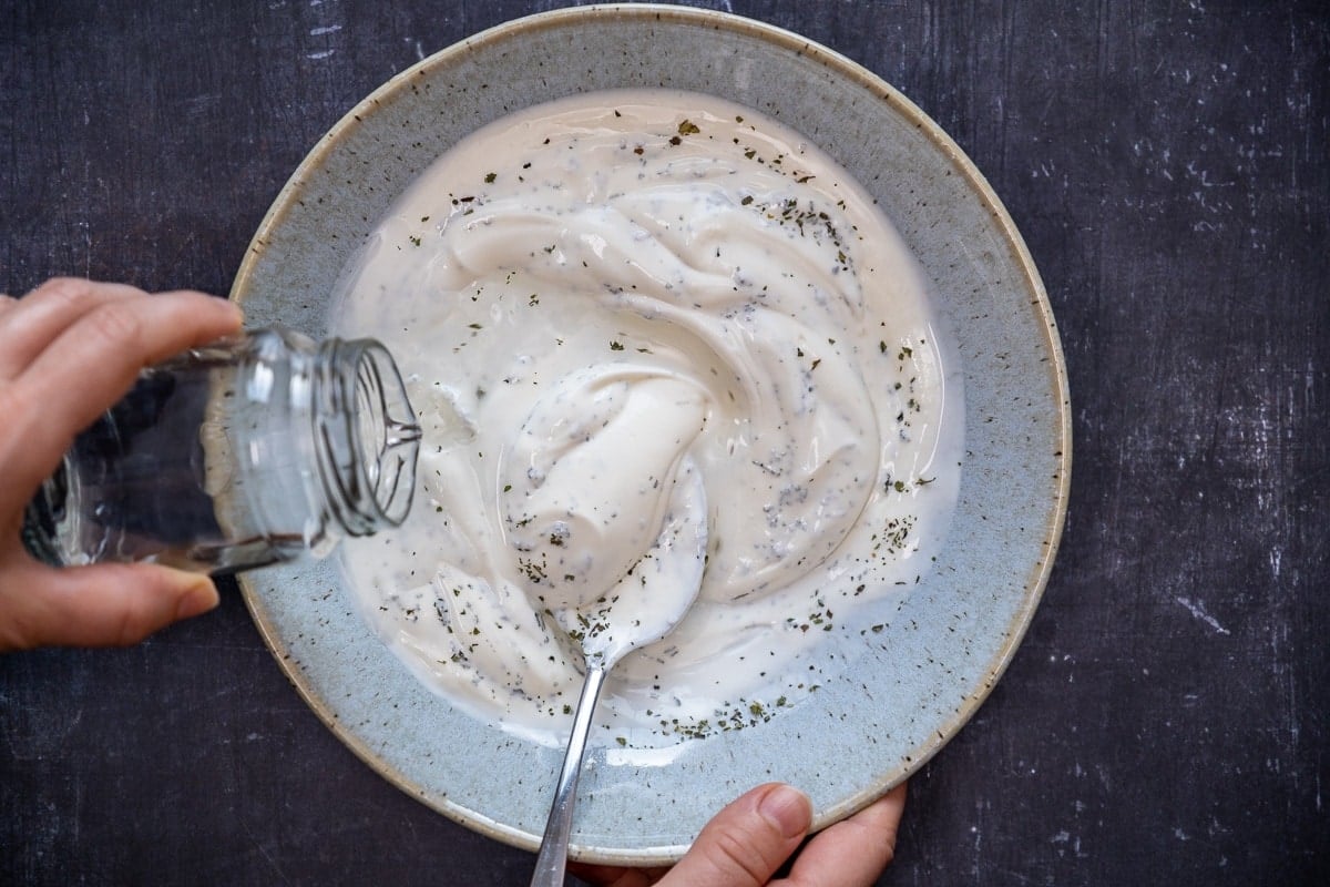 Hands pouring water from a small glass bottle into the mixture of yogurt, dried mint and salt in a blueish bowl. A spoon inside it.