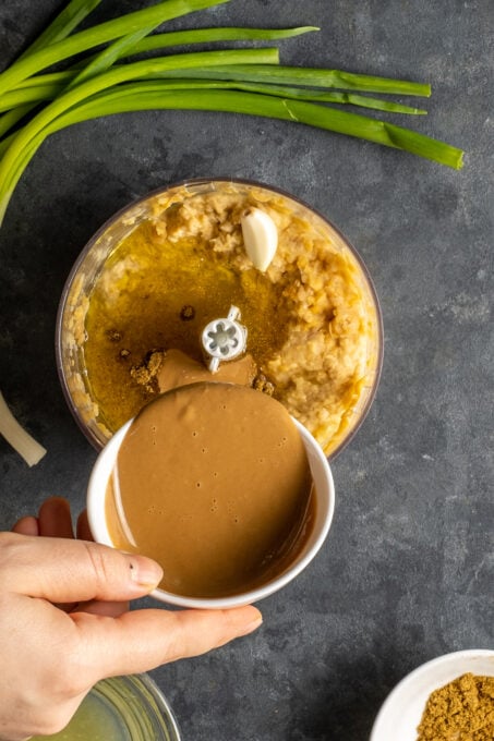 A hand pouring tahini from a small white bowl on mashed chickpeas in a food processor.