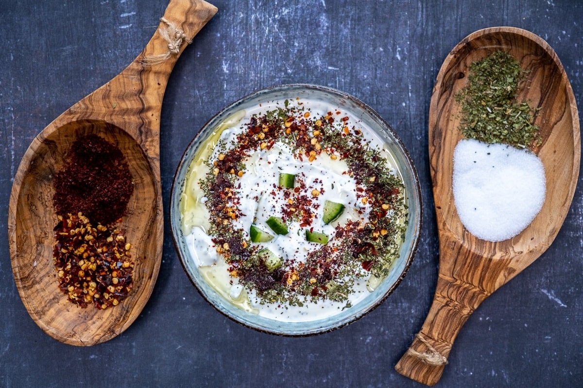 Turkish cacik topped with olive oil, dried mint, sumac and red pepper flakes in a bowl and two wooden bowls with spices on both sides of the bowl on a dark background.