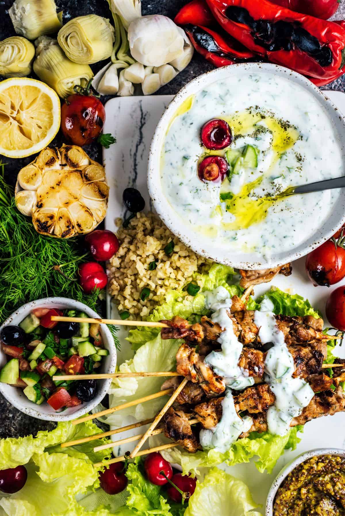 Yogurt cucumber cacik in a white bowl accompanied by chicken kebabs, shepherd salad, lettuce leaves, roasted garlic, artichoke hearts, roasted red peppers, bulgur pilaf, tomatoes and cherries.