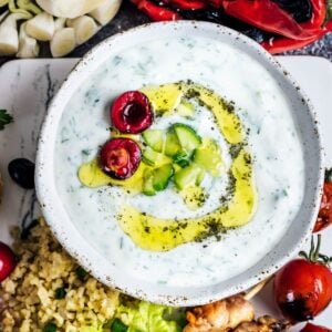 Yogurt and cucumber cacik in a white ceramic bowl, drizzled with olive oil and topped with chopped cucumber and halved cherry.