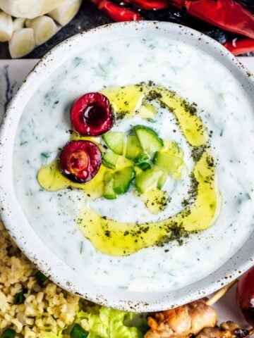 Yogurt and cucumber cacik in a white ceramic bowl, drizzled with olive oil and topped with chopped cucumber and halved cherry.