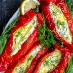 Feta stuffed roasted red peppers served on a plate, garnished with dill and lemon, with garlic cloves in the background.
