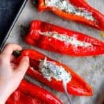 Hands filling roasted red peppers with a herbed feta cheese mixture on a baking tray lined with parchment paper.