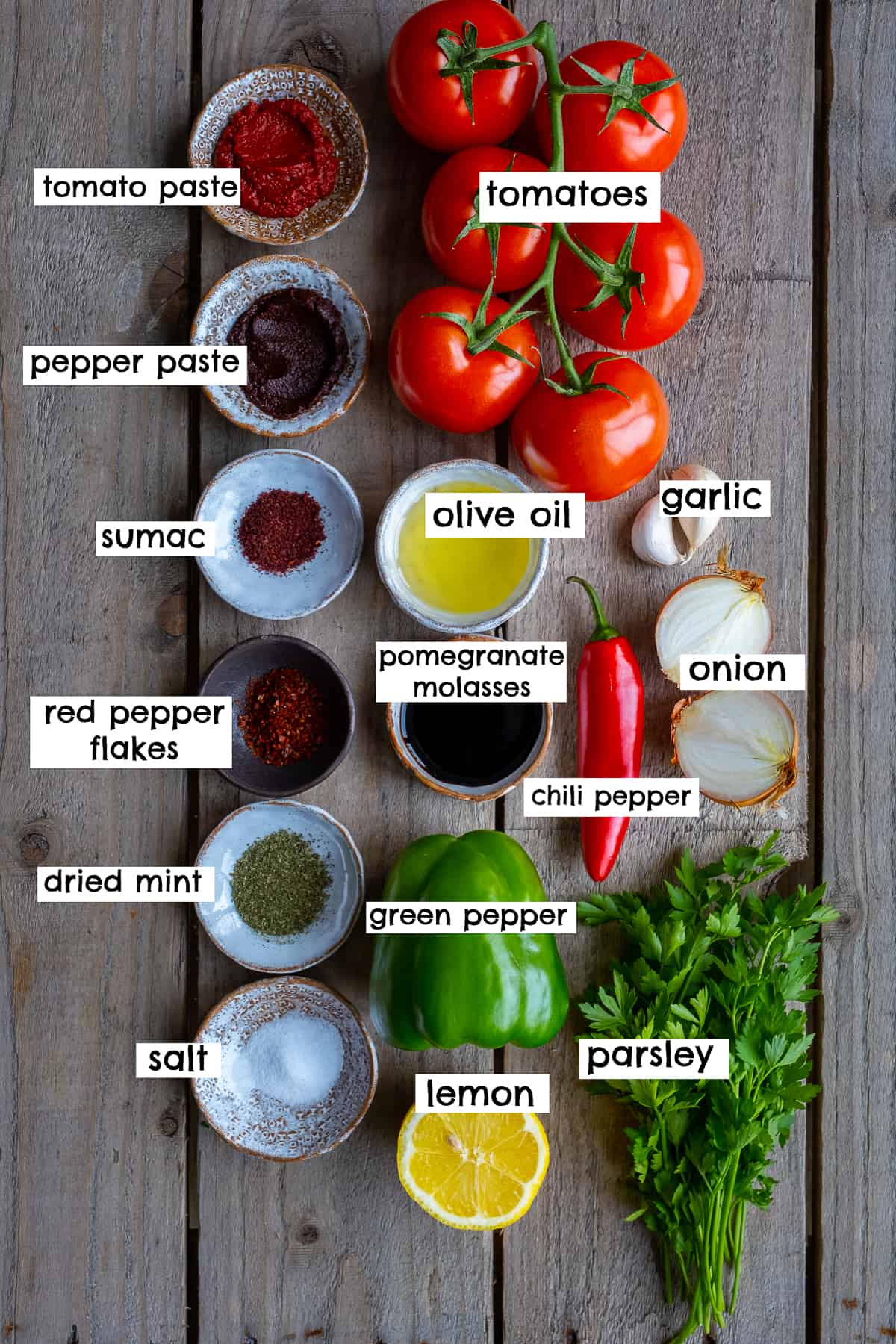 Spices, tomato paste, pepper paste, tomatoes, olive oil, lemon, parsley, red chili pepper, green bell pepper, halved onion, garlic cloves, pomegranate molasses on a wooden backdrop.