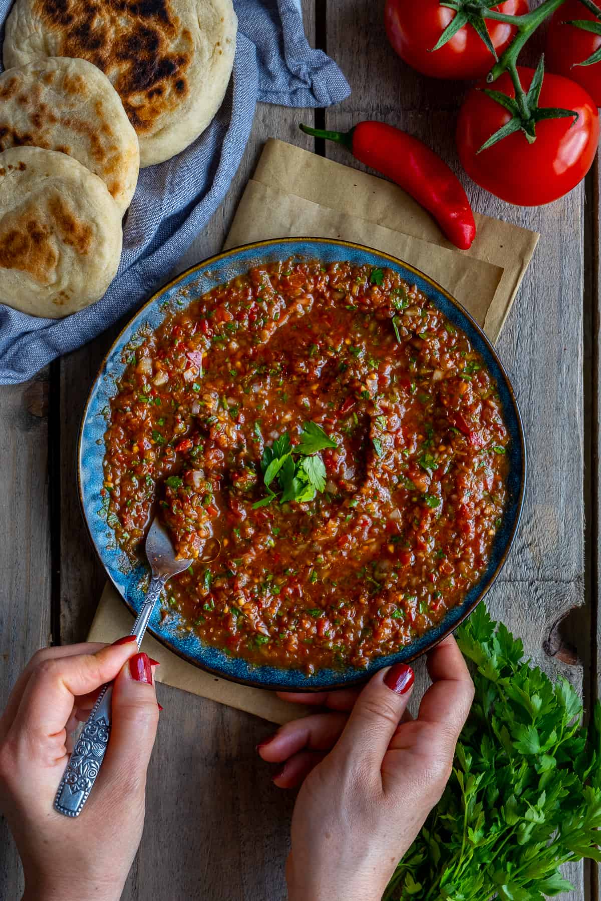 Hands holding a plate loaded with ezme dip and taking some of it with a fork, a red chili pepper, tomatoes, parsley and flatbreads on the side.