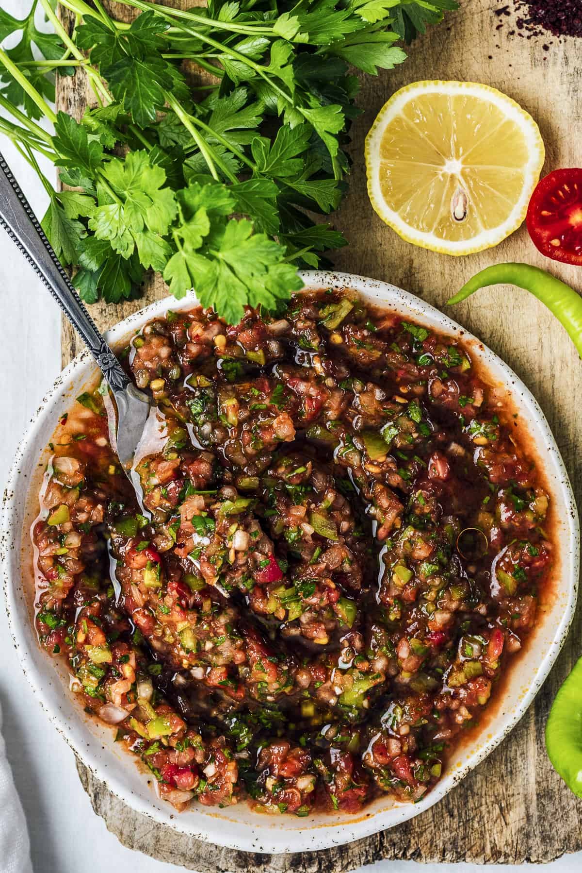 Turkish ezme salad on a white ceramic plate with a fork inside it. A bunch of parsley and a slice of lemon on the side.