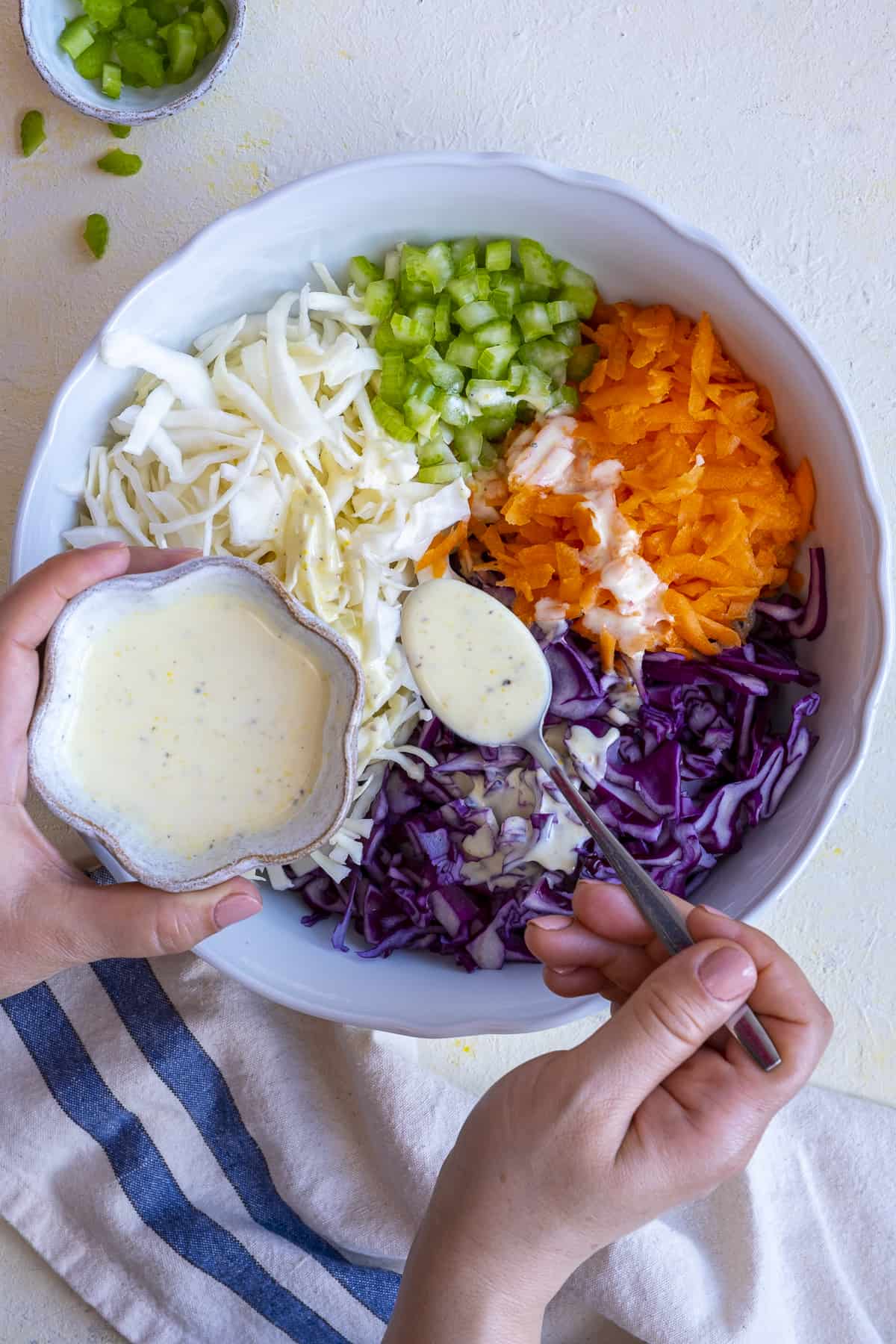 Hands pouring yogurt dressing over coleslaw base mixture in a bowl.