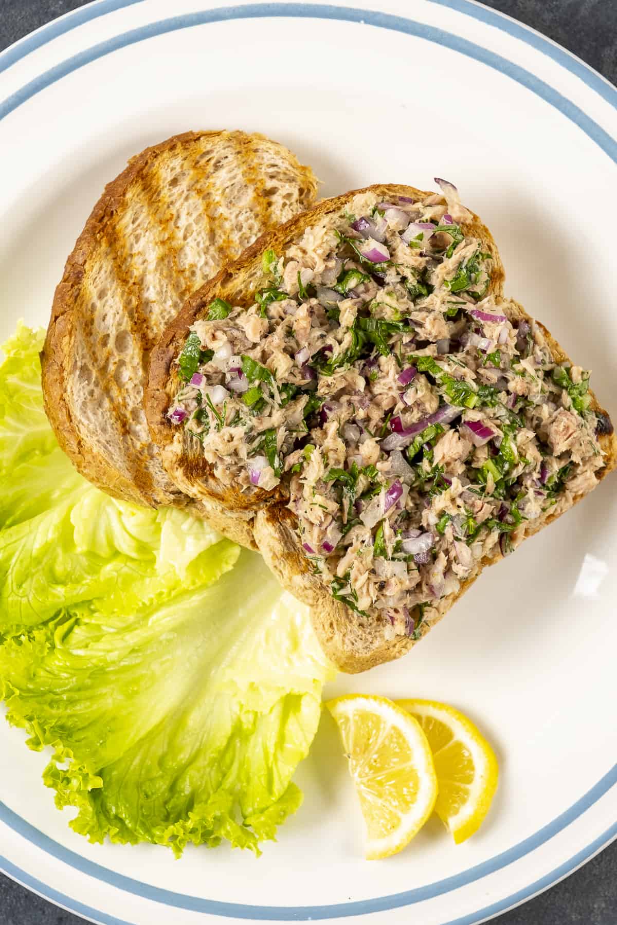 Tuna salad served on a toasted bread slice and accompanied by lettuce leave and lemon wedges.