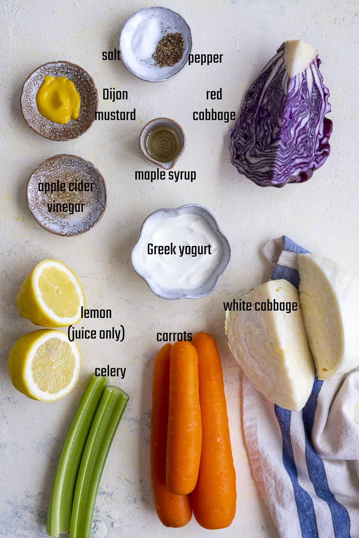 Red cabbage, white cabbage, carrots, lemon, celery ribs, Greek yogurt, Dijon mustard, vinegar, maple syrup, salt and pepper on a light background.