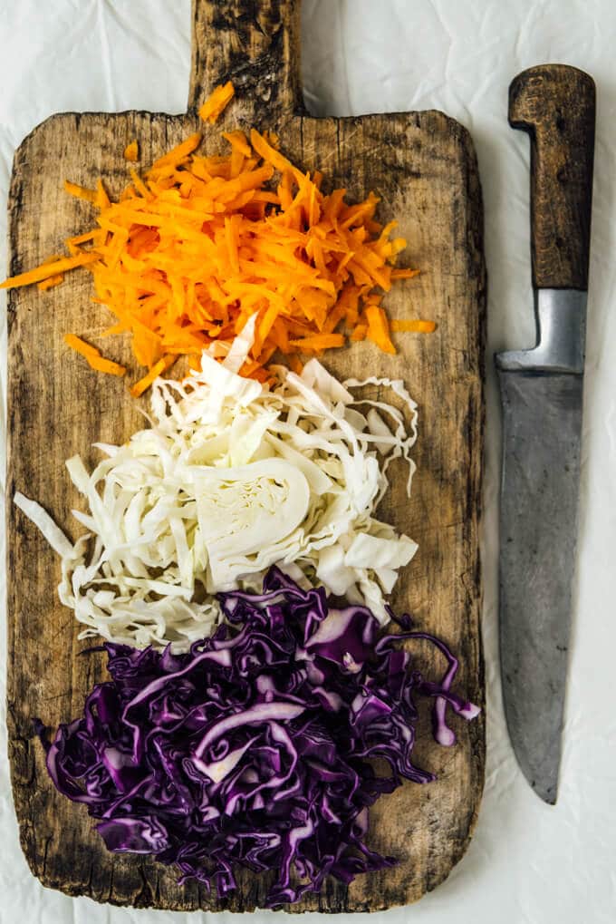 Shredded red cabbage, white cabbage and carrots on a wooden cutting board and a knife on the side.