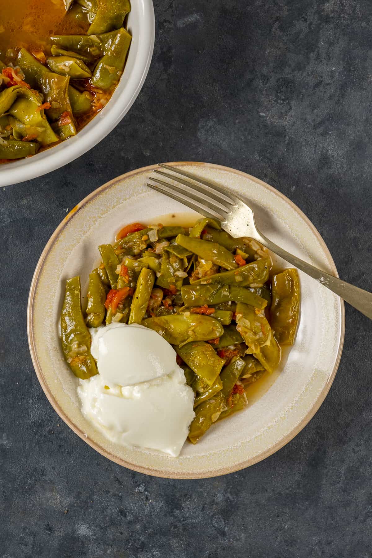 Turkish style green beans served in a white bowl with some yogurt on the side pictured on a dark background.