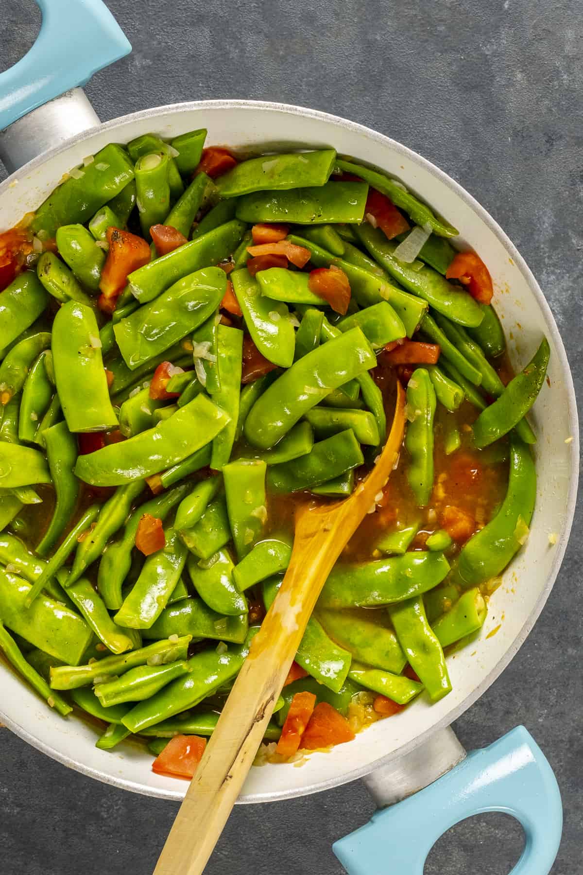 Chopped green beans and tomatoes cooking in a pot and a wooden spoon inside it.