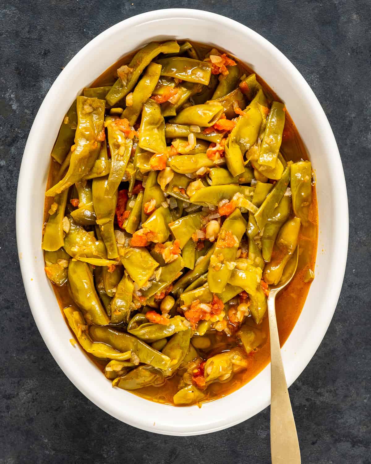 Turkish-style green beans cooked in olive oil with tomatoes and onions, served in an oval white dish with a spoon.