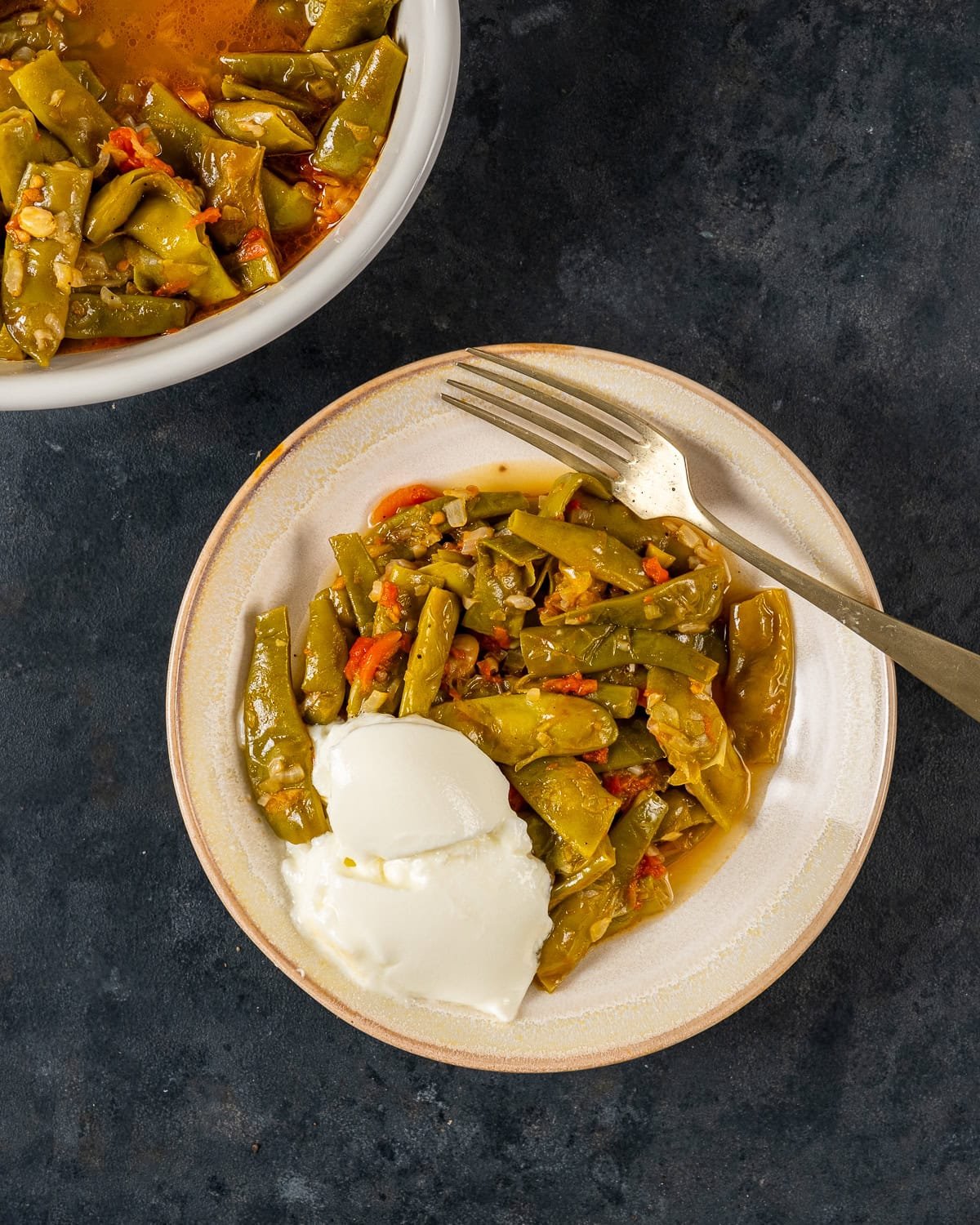 A serving of Turkish green beans with a side of yogurt on a plate, with a fork.