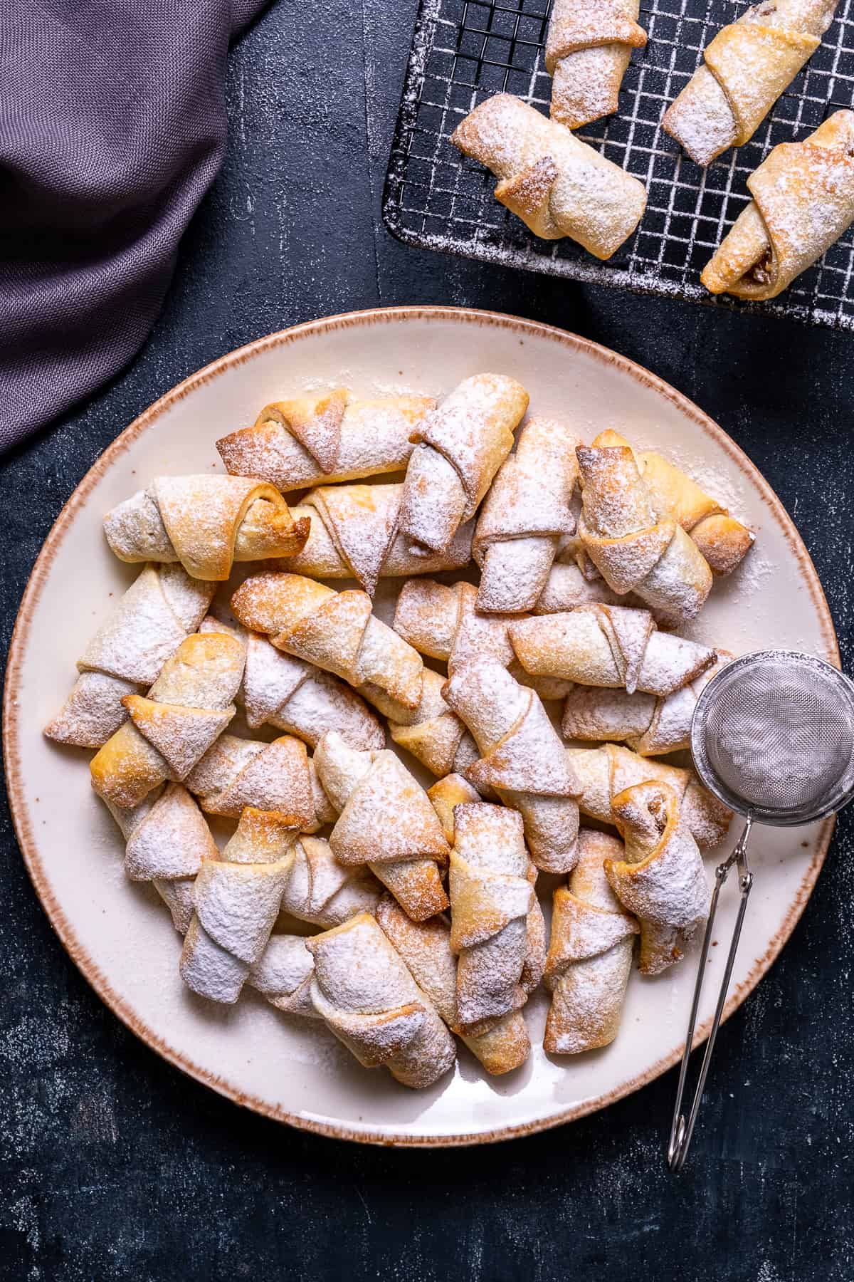 Turkish cookies on a white plate, powdered sugar sprinkled on them, some more cookies on a cooling rack on the side.