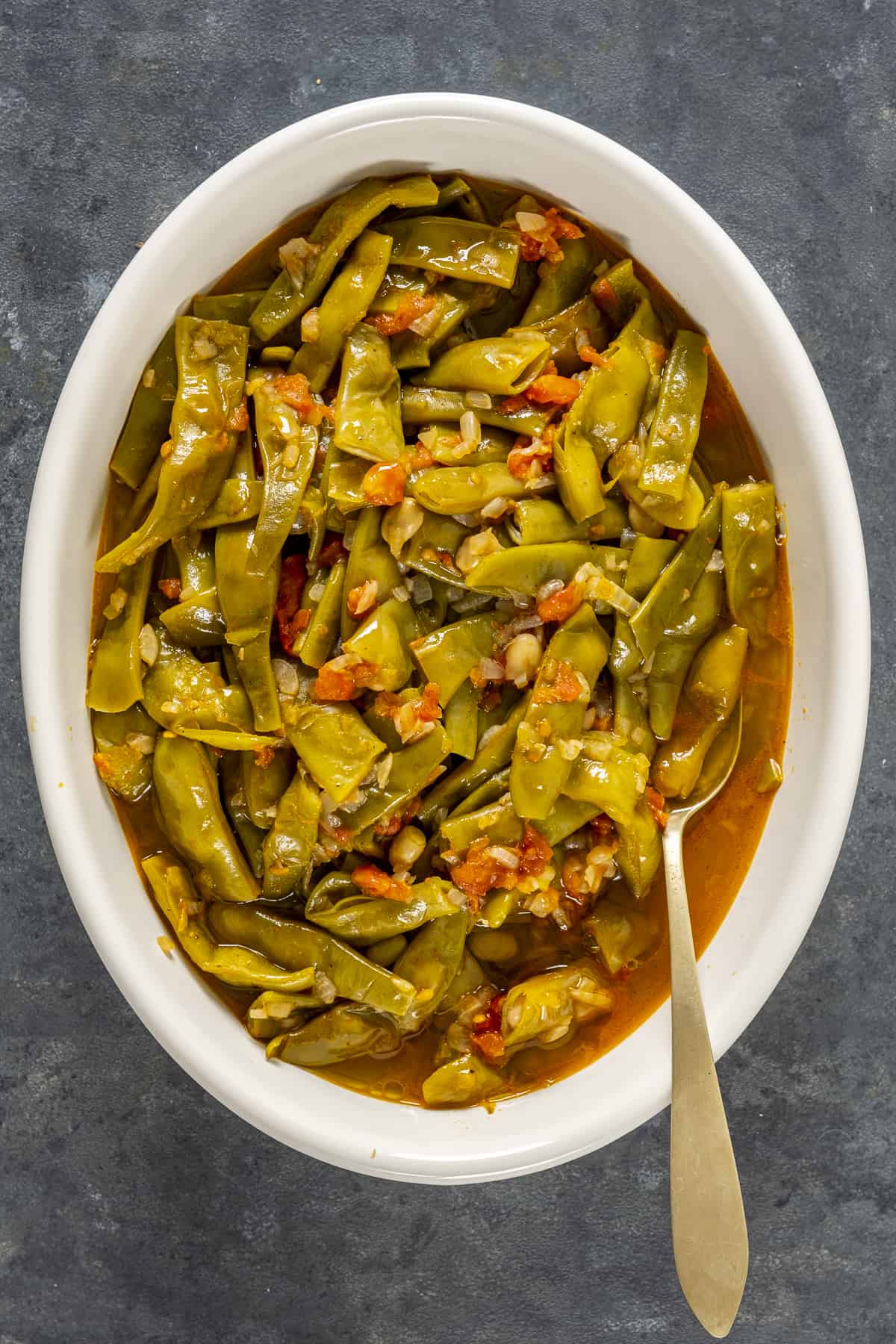 Green beans with tomatoes served in a white oval dish and a spoon inside it.