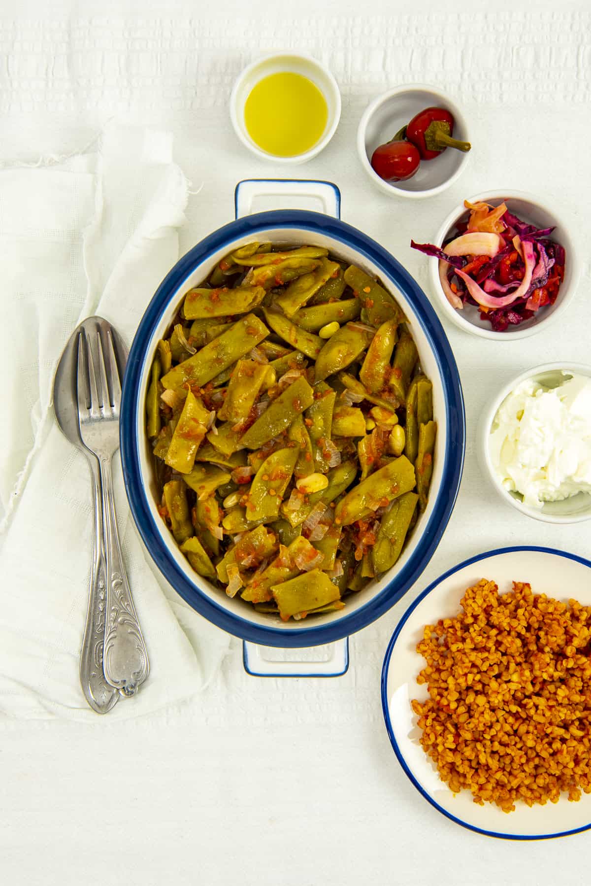 Turkish green beans served in a white oval dish and bulgur pilaf, pickles and yogurt on the side.