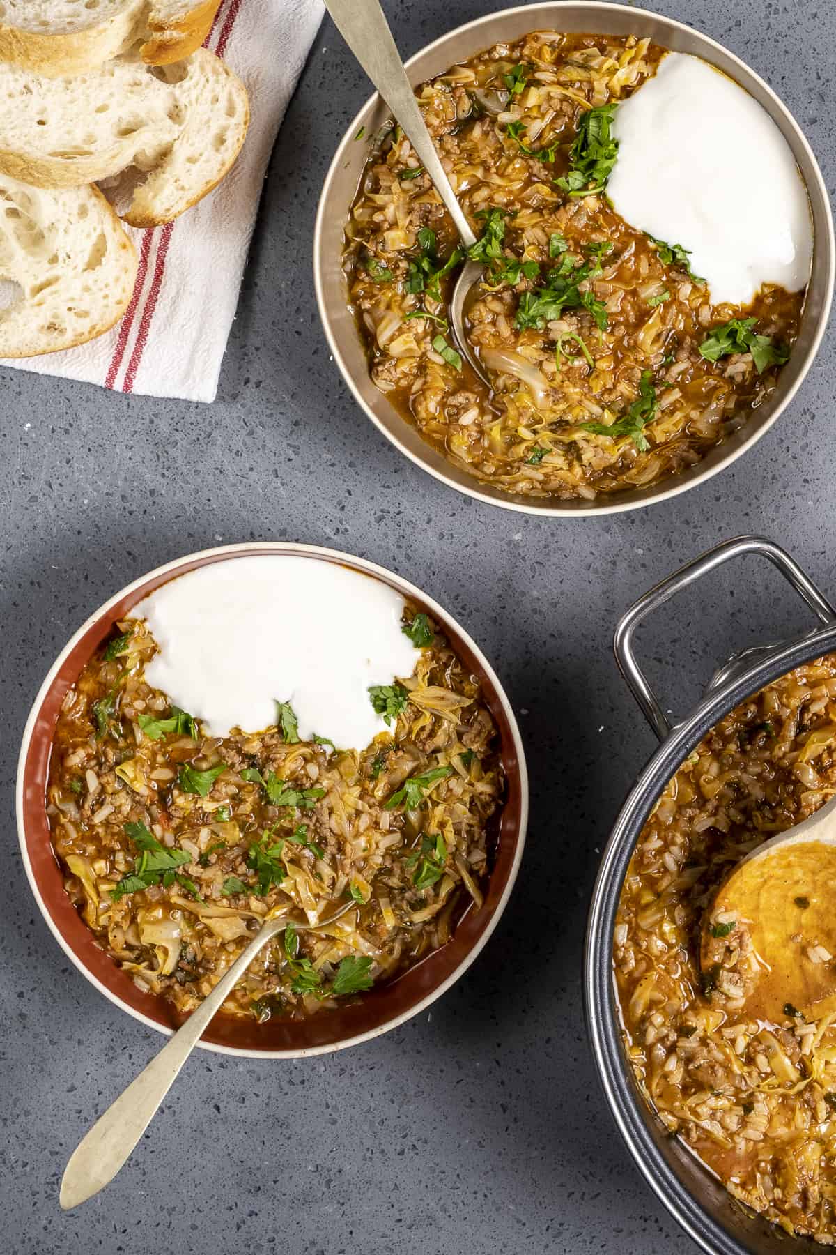 Stuffed cabbage soup in two bowls garnished with yogurt, some bread and the pot on the side.