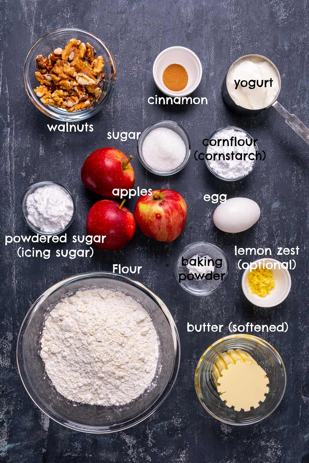 Apples, walnuts, sugar, egg, lemon zest, butter, flour, cinnamon, icing sugar and baking powder on a dark background.