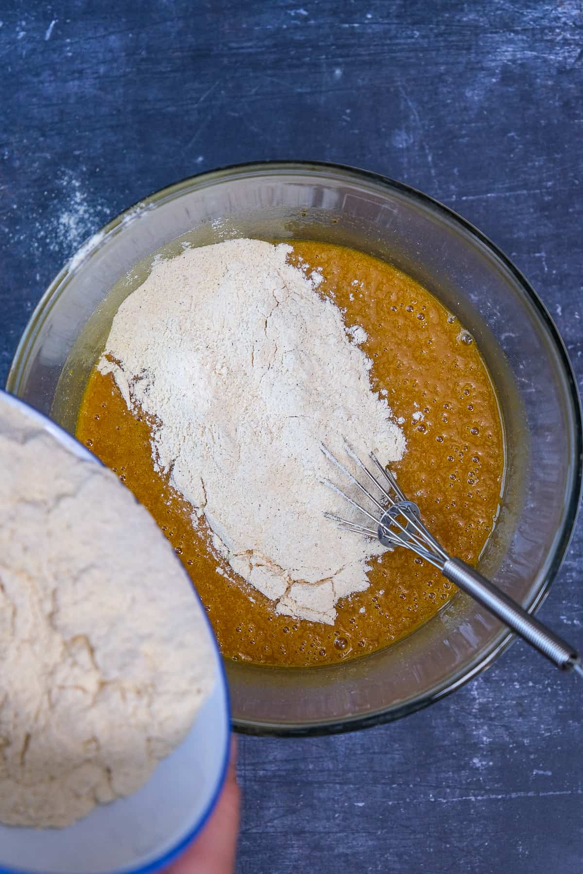 Adding dry ingredients from a white bowl into the wet mixture in a glass bowl.