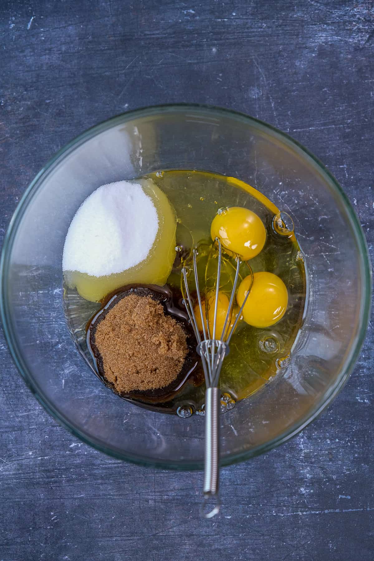 Eggs and dark and granulated sugars in a glass mixing bowl with a hand whisk inside it.