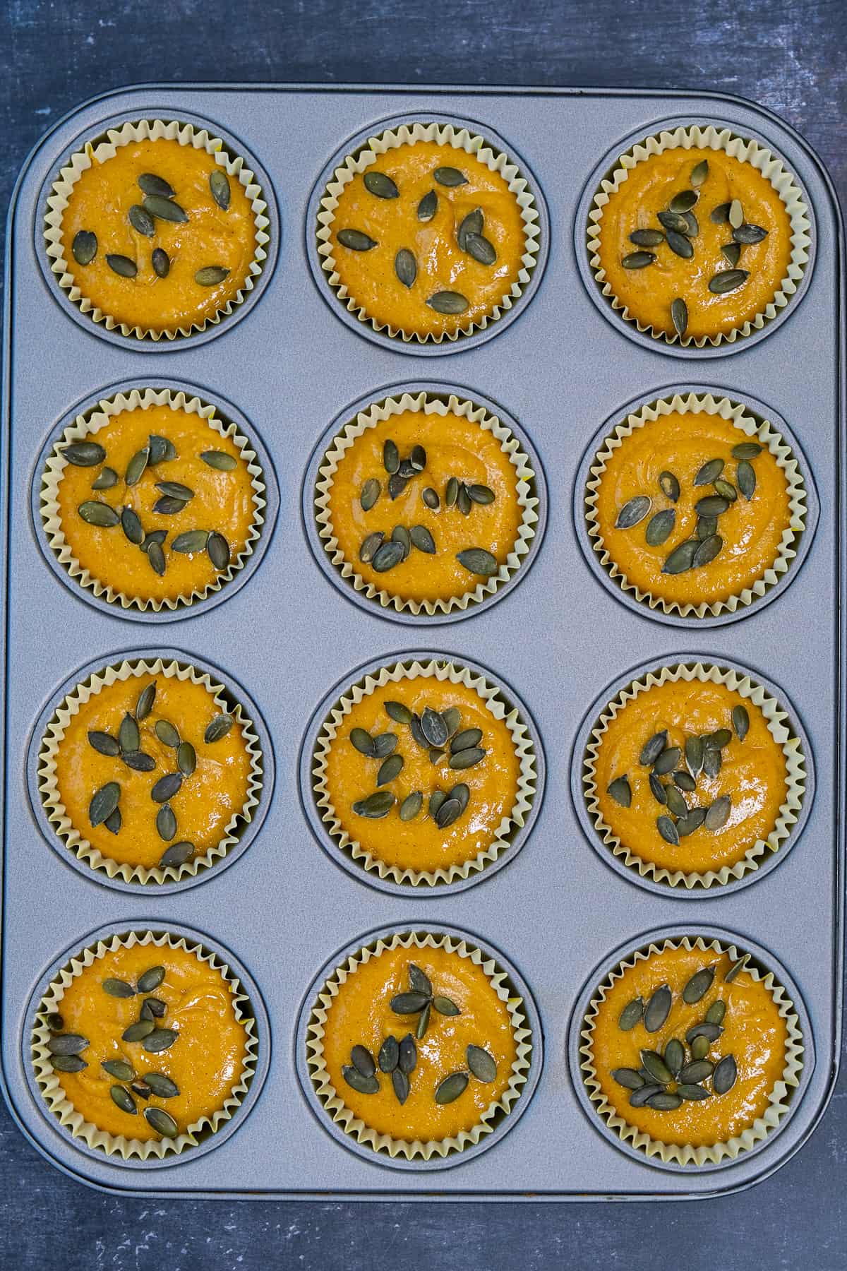 Pumpkin muffin batter shared into a muffin pan lined with muffin liners and topped with pumpkin seeds.