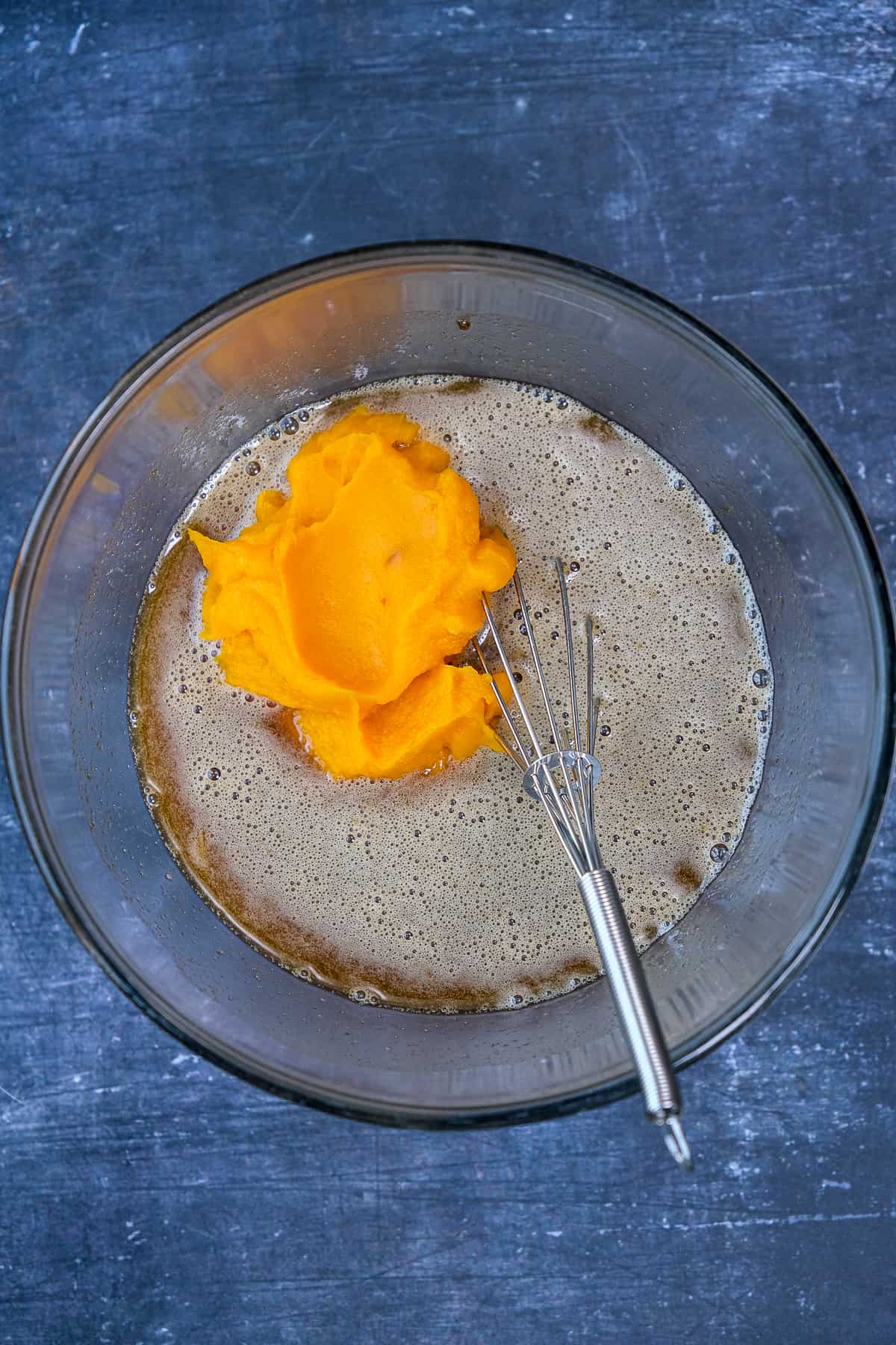 Eggs and sugars are creamed and pumpkin puree added in a glass mixing bowl. A hand whisk inside it.