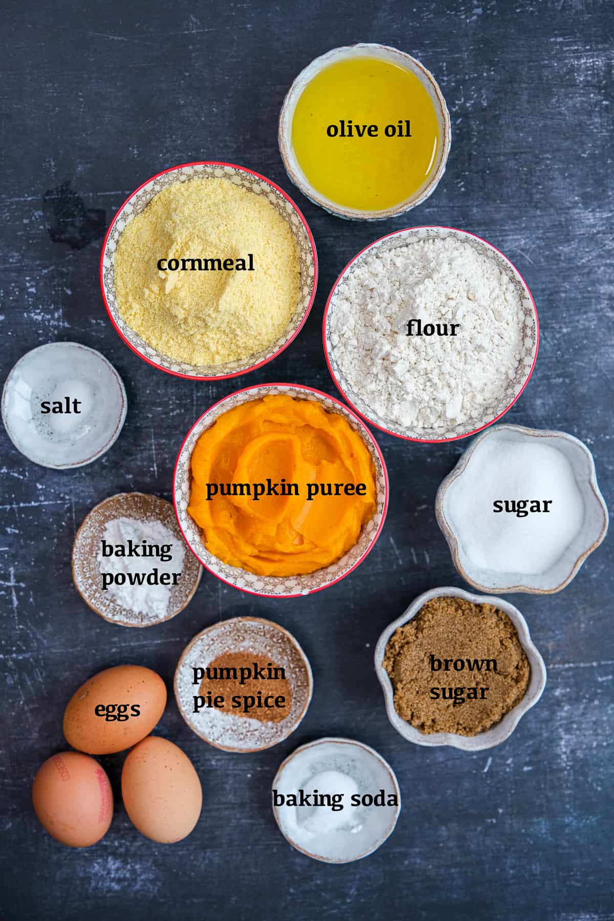 Pumpkin puree, cornmeal, flour, olive oil, brown sugar, granulated sugar, eggs, pumpkin spice, salt, baking soda and baking powder all in separate bowls on a dark background.