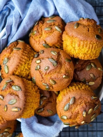 Pumpkin muffins topped with pepitas in a white bowl lined with blue napkin.