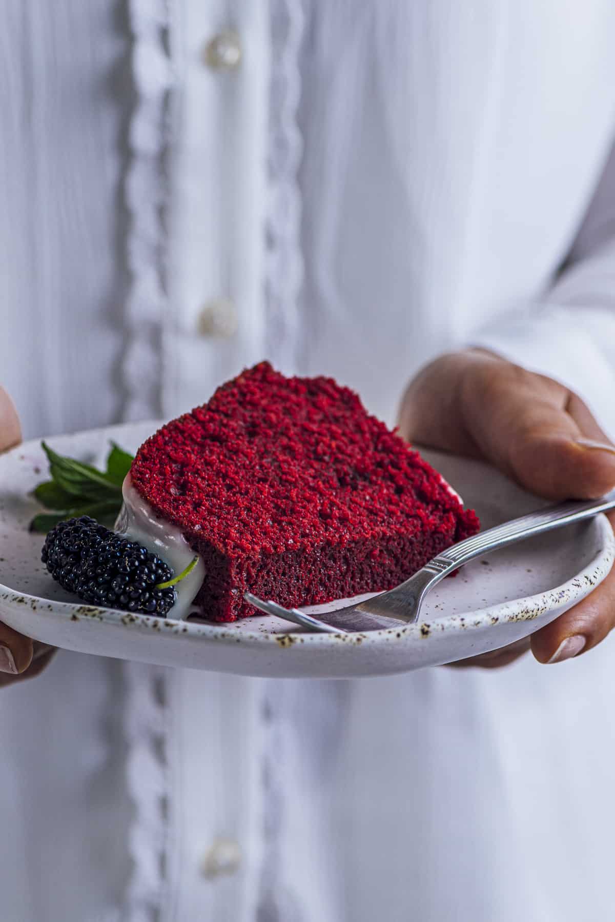 Woman with a white blouse holding a slice of red velvet bundt cake on a white plate.