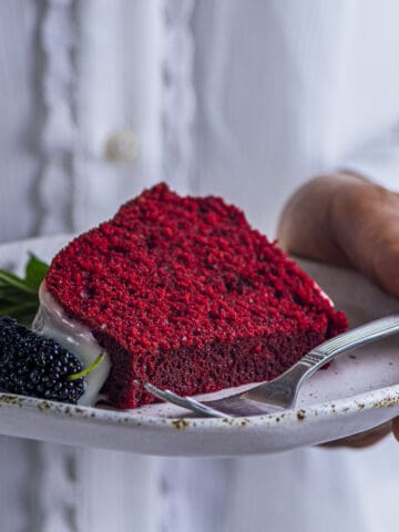 A woman with white shirt holding a slice of red velvet bundt cake with white glaze, a mulberry and mint garnish in a ceramic white plate.