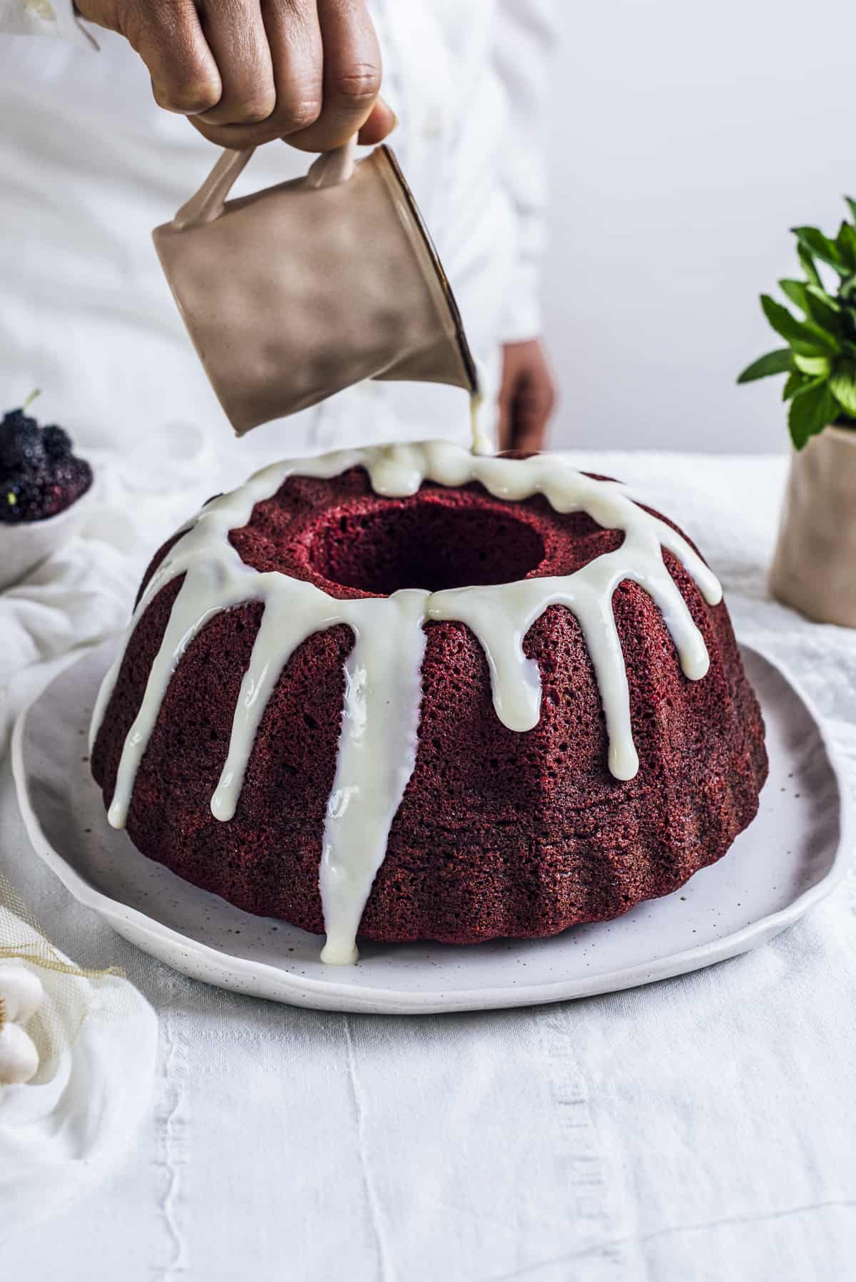 A woman with a white shirt pouring a white glaze on a red velvet bundt cake photographed from front view.