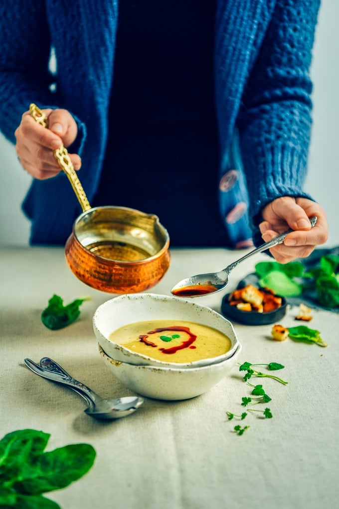 Woman drizzling chili oil sauce from a copper saucepan over spicy red lentil soup in a white bowl.