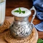 A traditional metal cup filled with frothy Turkish ayran and garnished with mint, shown with text overlay that reads “Ayran – Turkish Yogurt Drink” and the website “giverecipe.com” at the bottom.