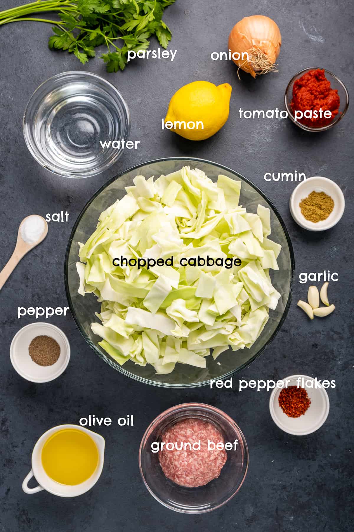 Chopped cabbage in a bowl, ground beef, olive oil, tomato paste, lemon, garlic cloves, onion, parsley, cumin, salt and pepper on a dark background.