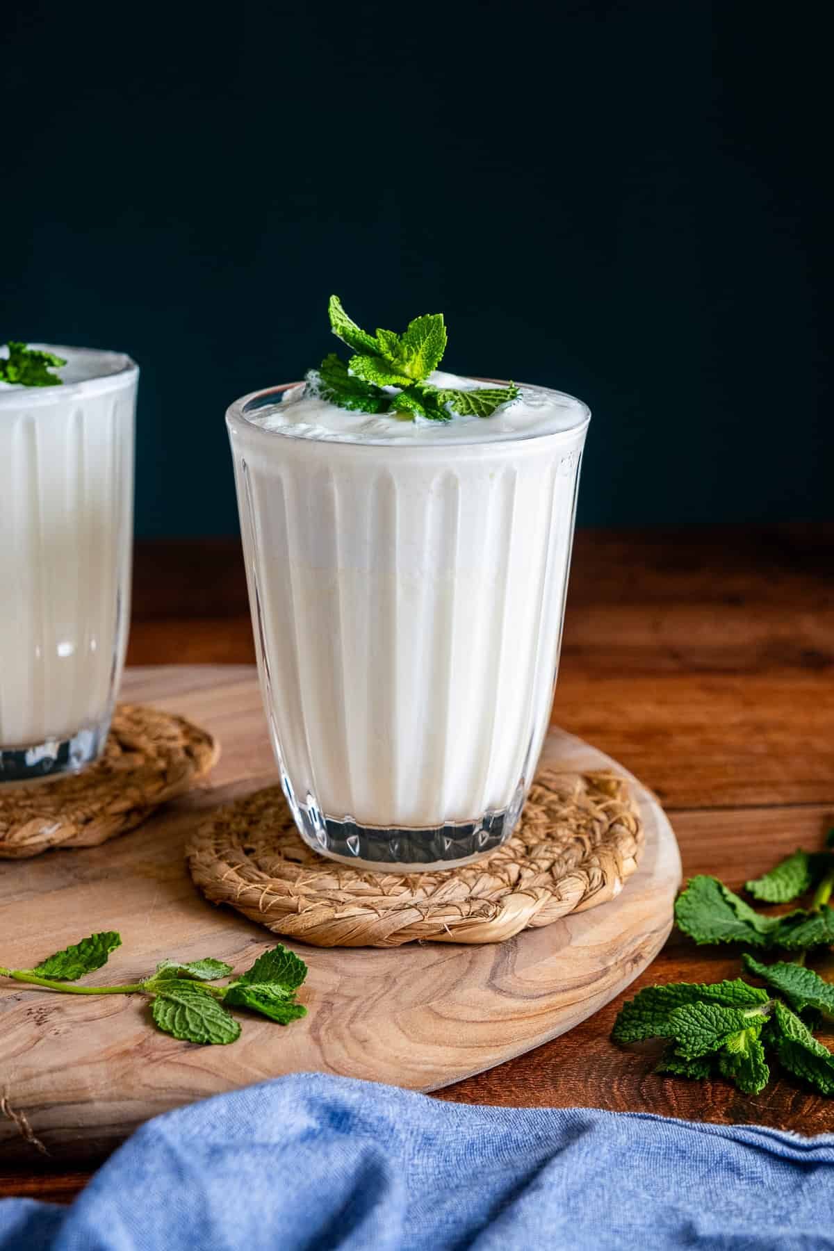 A glass of foamy Turkish ayran topped with fresh mint, placed on a woven coaster over a wooden board, with mint leaves and a blue cloth nearby.