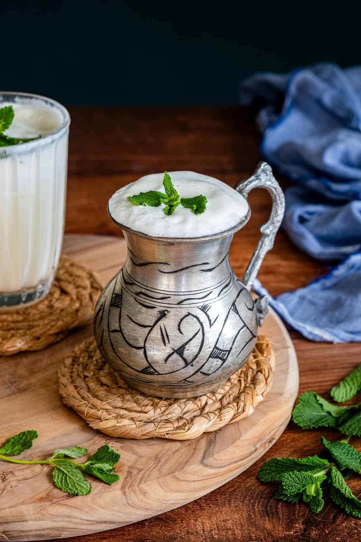 A traditional metal cup filled with frothy Turkish ayran, garnished with fresh mint, placed on a woven coaster with a wooden board and mint leaves around it.