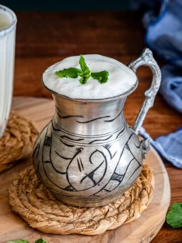 A traditional metal cup filled with frothy Turkish ayran, garnished with fresh mint, placed on a woven coaster with a wooden board and mint leaves around it.