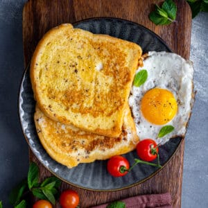Two slices of eggy bread served with tomatoes and fried egg garnished with mint leaves on a dark colored plate.