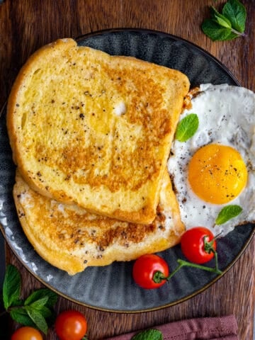 Two slices of eggy bread served with tomatoes and fried egg garnished with mint leaves on a dark colored plate.