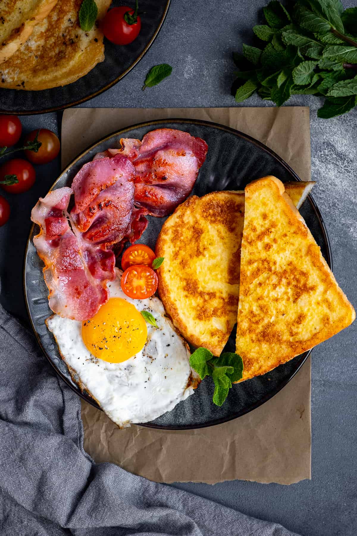 Eggy bread sliced in half served with bacon, fried egg and tomatoes on a dark plate.