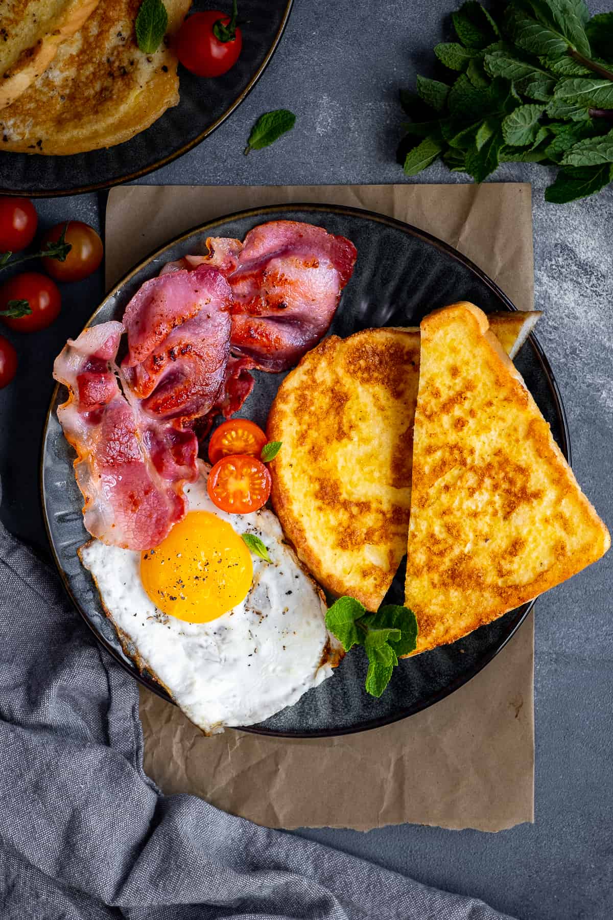 Eggy bread sliced in half served with bacon, fried egg and tomatoes on a dark plate.