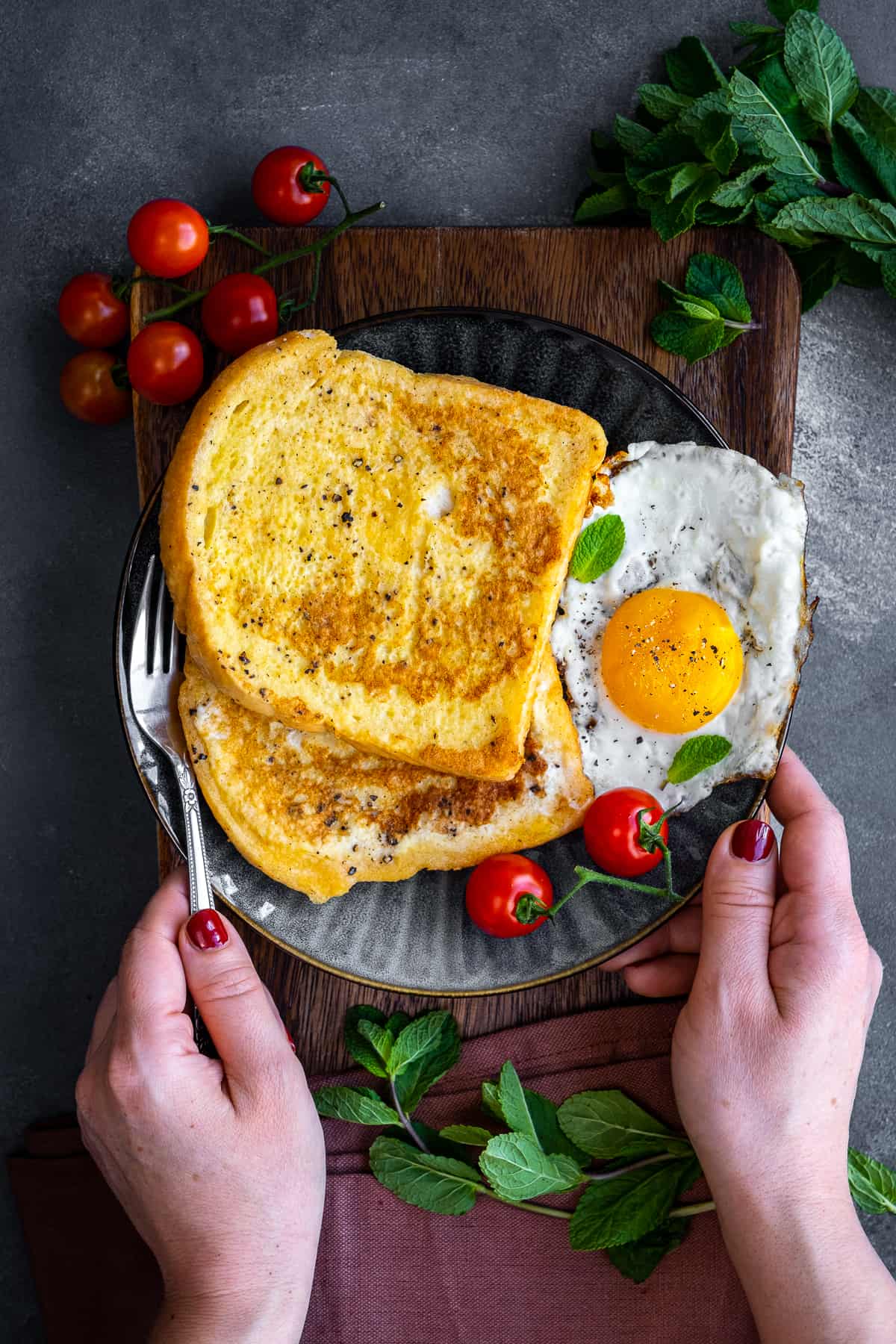 Woman about to eat eggy bread served with fried egg and cherry tomatoes on a grey colored plate.