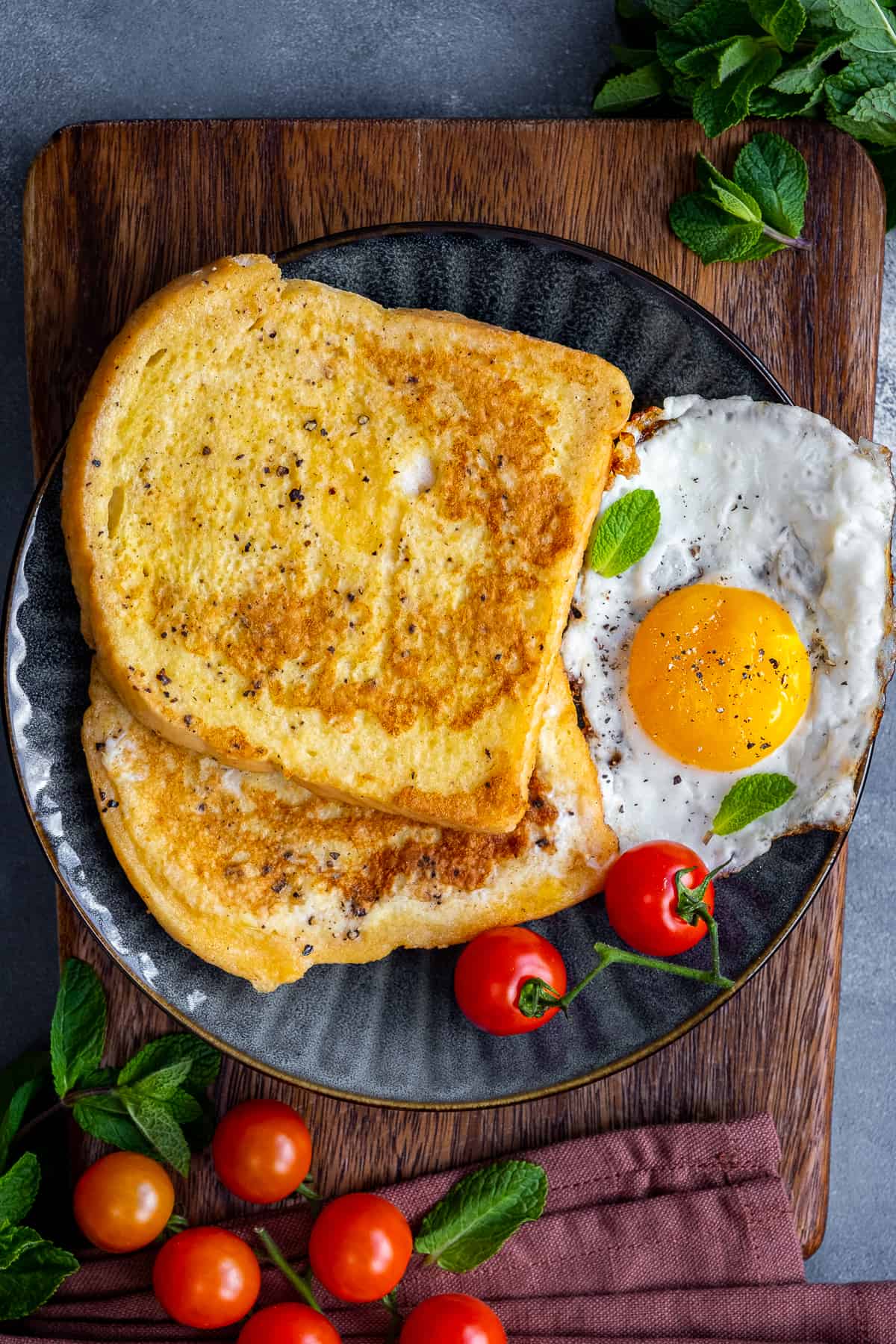 Eggy bread served with fried egg and tomatoes on a dark colored plate. Cherry tomatoes and fresh mint leaves on the side.