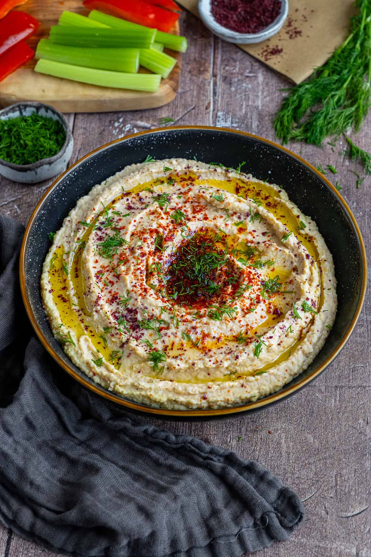 Hummus topped with spices and fresh dill in a black bowl and celery sticks and red bell sticks behind it.
