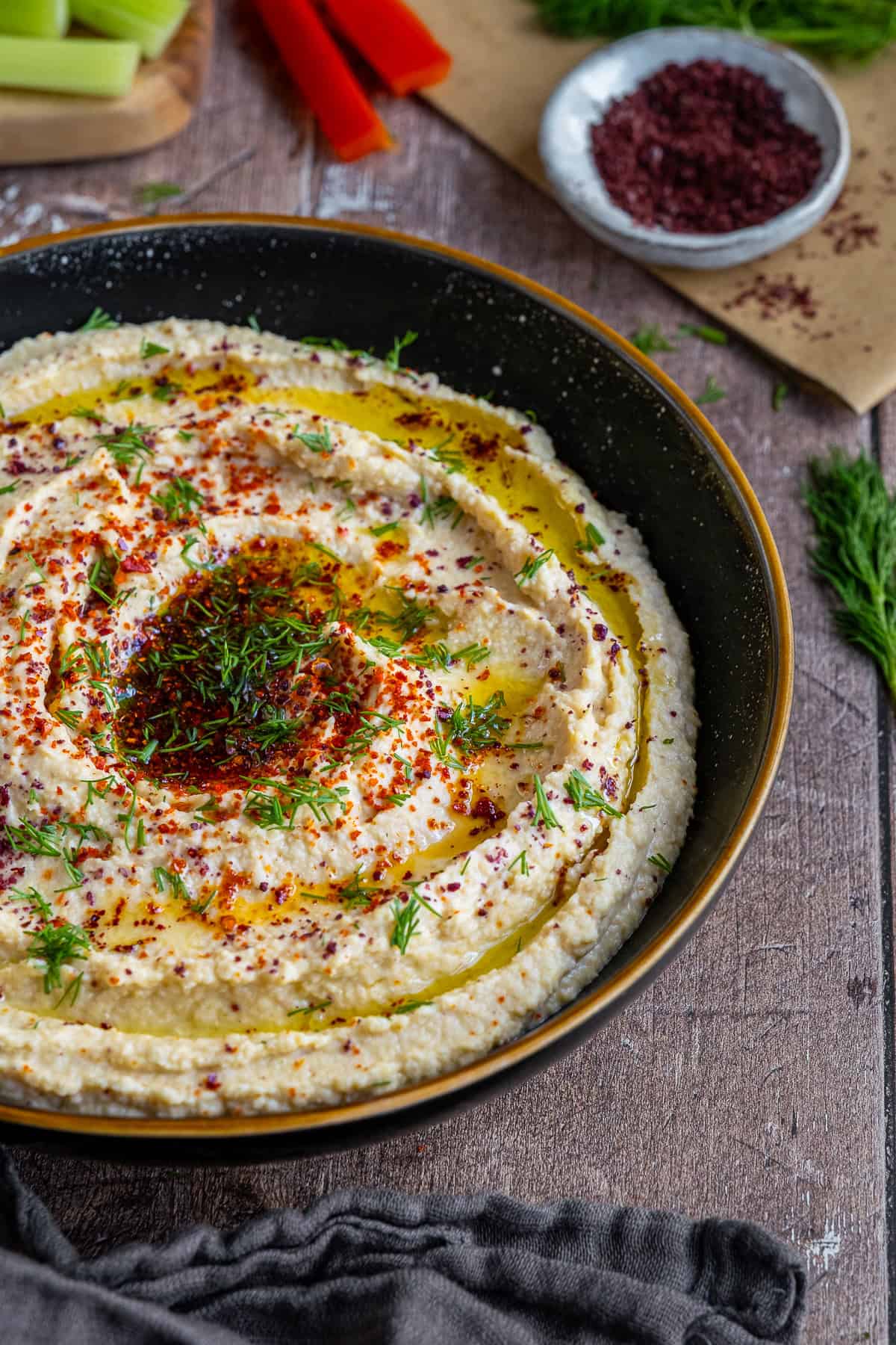 Hummus garnished with red pepper flakes, sumac and fresh dill served in a black bowl.